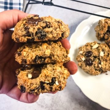 three oatmeal cookies held in hand with plated cookies on a white plate and black cooling rack in the background