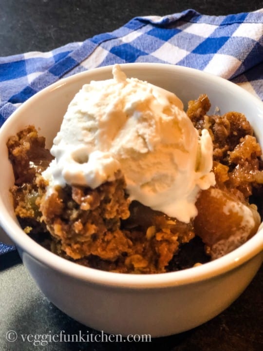 Apple spice dump cake in white bowl topped with ice cream with blue checkered napkin in background.