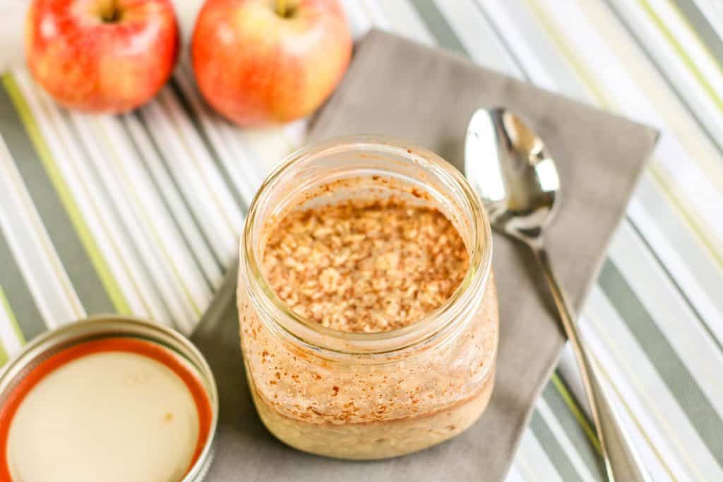 overnight oats with apple and cinnamon in jar with spoon and two apples on green striped placemat