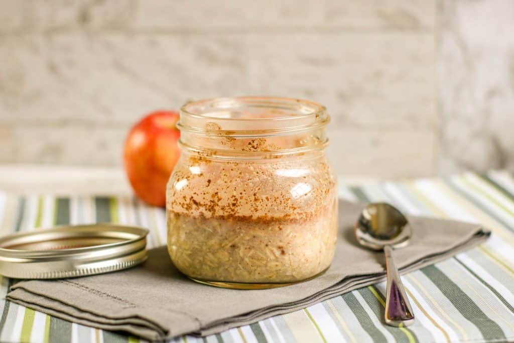 overnight oats with apple and cinnamon in jar with spoon and two apples on green striped placemat