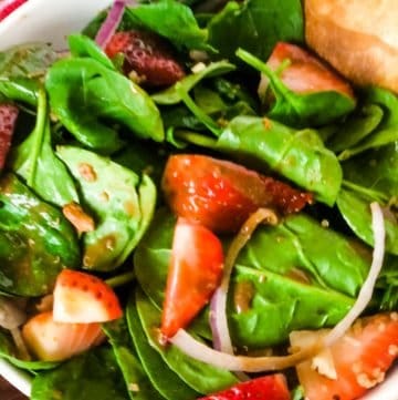 strawberry spinach salad in white bowl with red check napkin in background and dinner roll at side of bowl