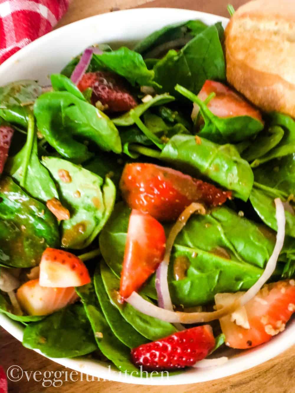 strawberry spinach salad in white bowl with red check napkin in background and dinner roll at side of bowl