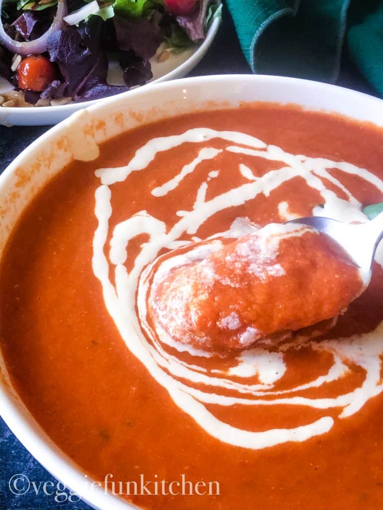 bowl of tomato soup with cashew cream drizzled and soup on spoon and green salad in background