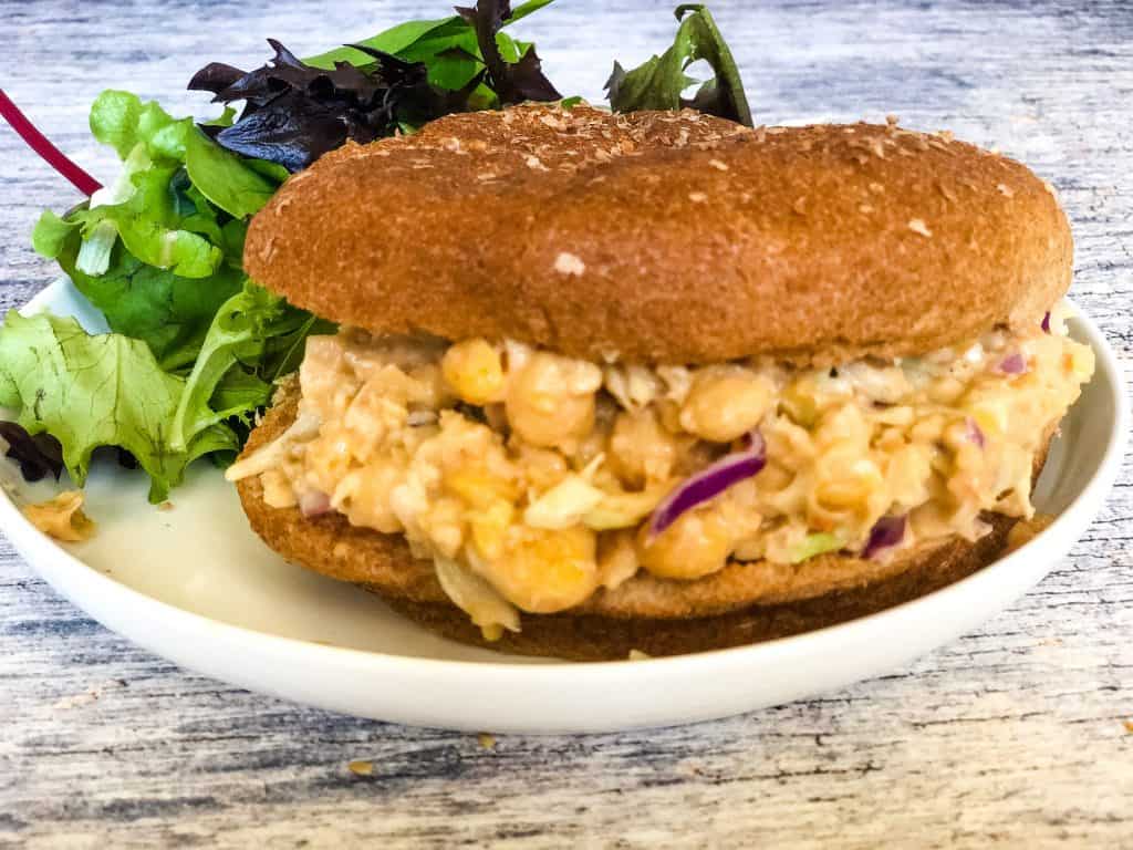 bbq chickpea salad sandwich on whole wheat bun on white plate with salad