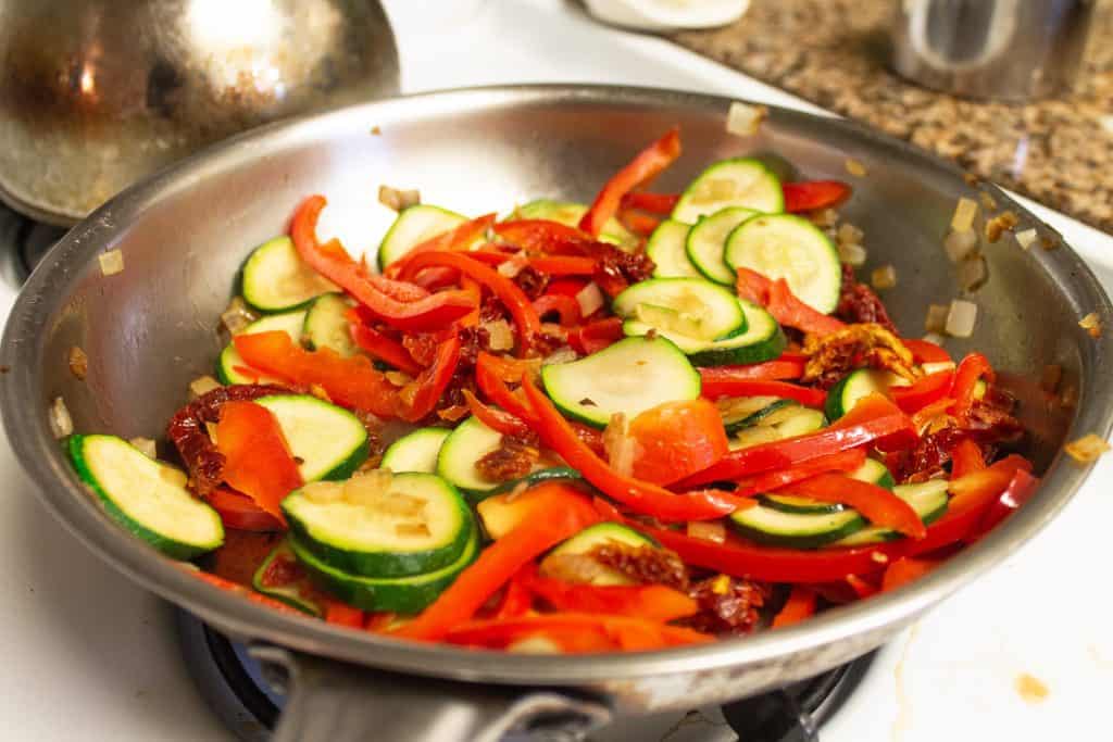 cut veggies including red bell pepper, zucchini, onion, and sun dried tomatoes in a stainless steel pan on stove-top