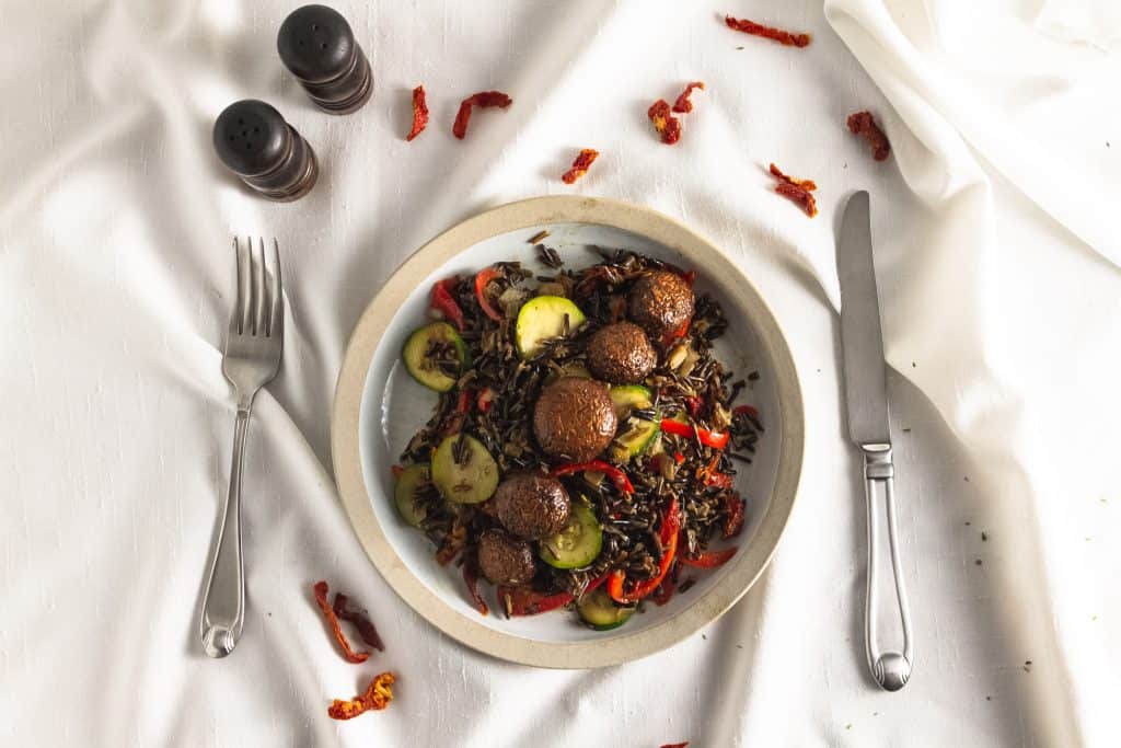Tan bowl of marinated mushrooms on a bed of wild rice and vegetables. on a white cloth with bits of sun-dried tomatoes scattered about