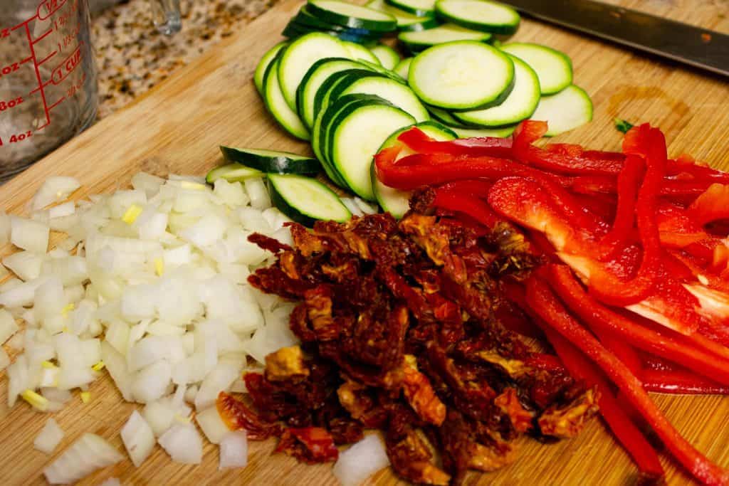 cut veggies on a wooden cutting board including onions, sun-dried tomatoes red bell peppers and zucchini - knife at side