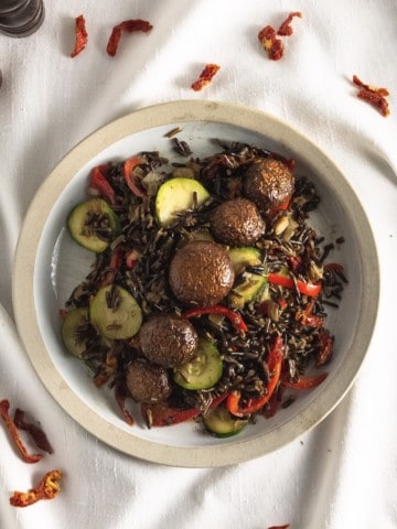 Tan bowl of marinated mushrooms on a bed of wild rice and vegetables. on a white cloth with bits of sun-dried tomatoes scattered about