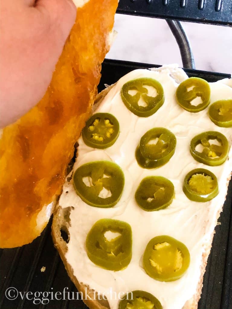 panini on press with vegan cream cheese and jalapeños on bottom layer with top layer being placed on top. Top later includes apricot jam.