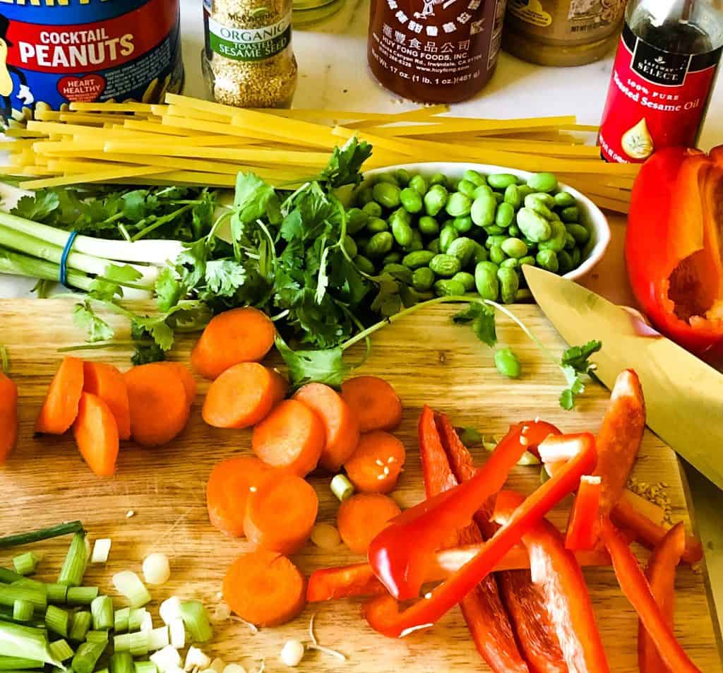 veggies for peanut butter noodles on brown cutting board including red peppers, sliced carrots, green onions, edamame, and cilantro. Noodles in background as well as some of the wet ingredients