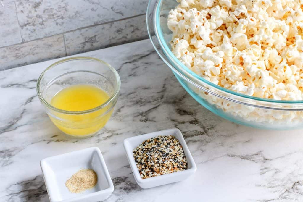 Ingredients for Everything Bagel popcorn including melted vegan butter, garlic powder, Everything Seasoning, and popped popcorn in a large glass bowl