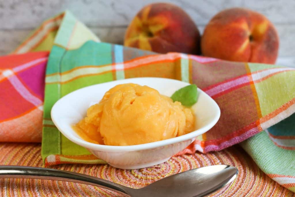 peach sorbet in white bowl on peach colored mat with spoon in front along with plaid napkin in back and two fresh peaches