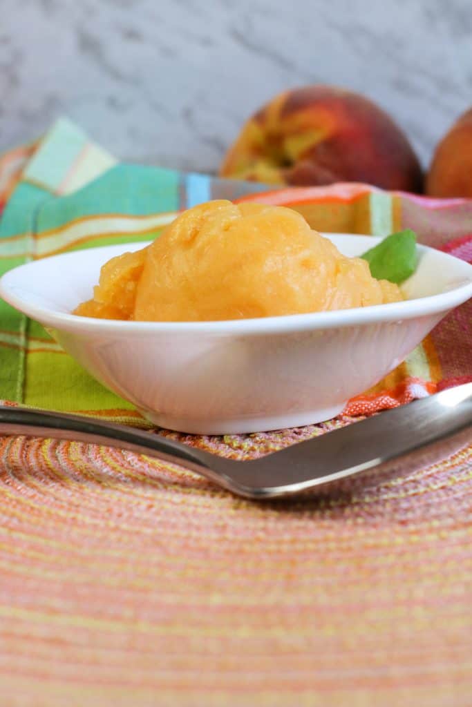 peach sorbet in white bowl on peach colored mat with spoon in front along with plaid napkin in back and two fresh peaches