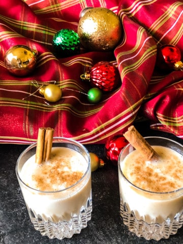 Two glasses of vegan egg-nog with cinnamon sticks against a red plaid background with christmas ornaments.