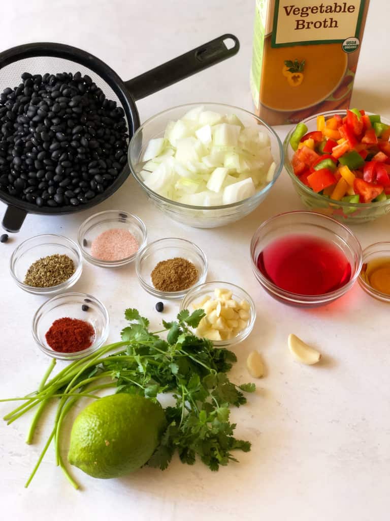 Ingredients for cuban black beans including dried beans, chopped onion bell peppers and onion, red wine vinegar, maple syrup, oregano, salt, cumin, smoked paprika, cilantro and lime.