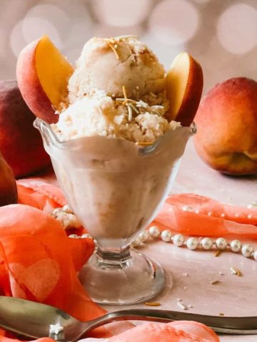 Peaches and cream ice cream in a glass dish with peaches sticking out of the top. a scarf, pearls, and spoon in the foreground. Peaches in the background.