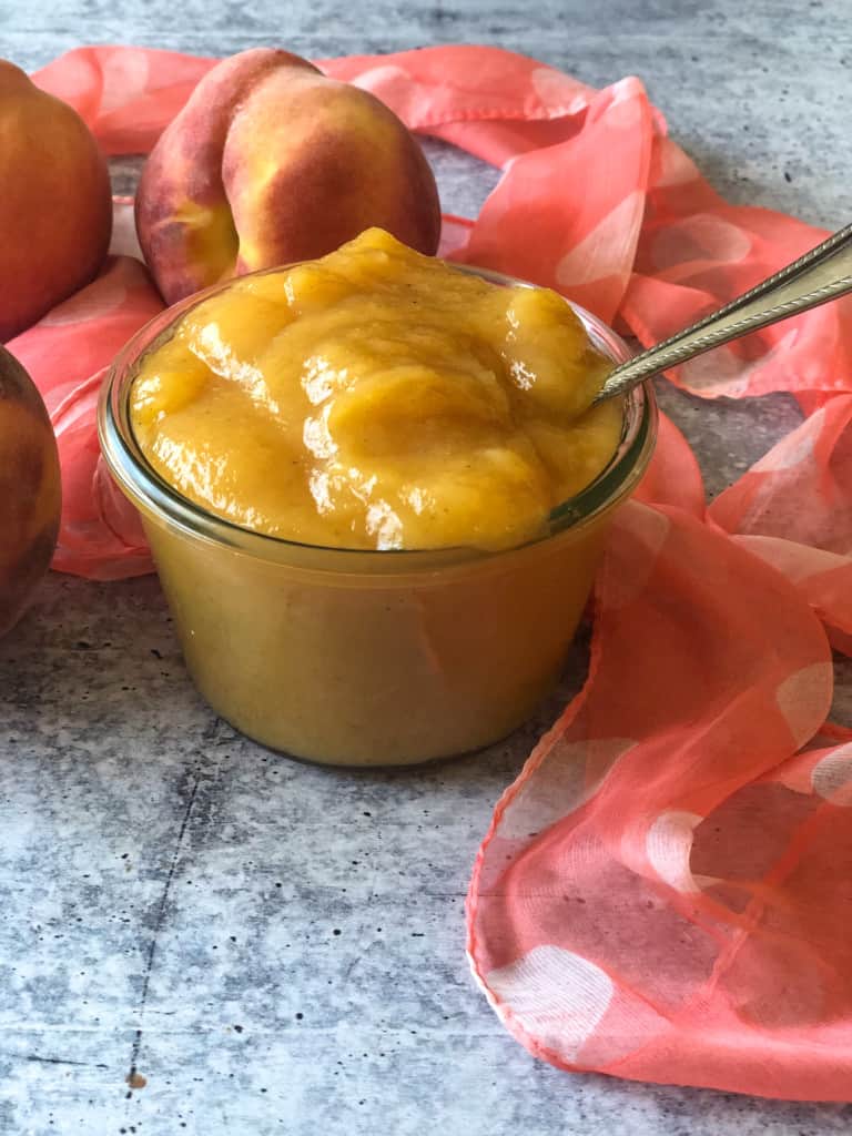 Peach butter in a glass container with peaches in background