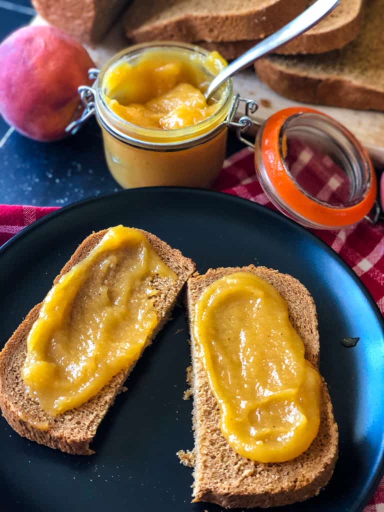 homemade wheat bread sliced on black plate and spread with peach butter - peaches, peach butter, and sliced bread in background