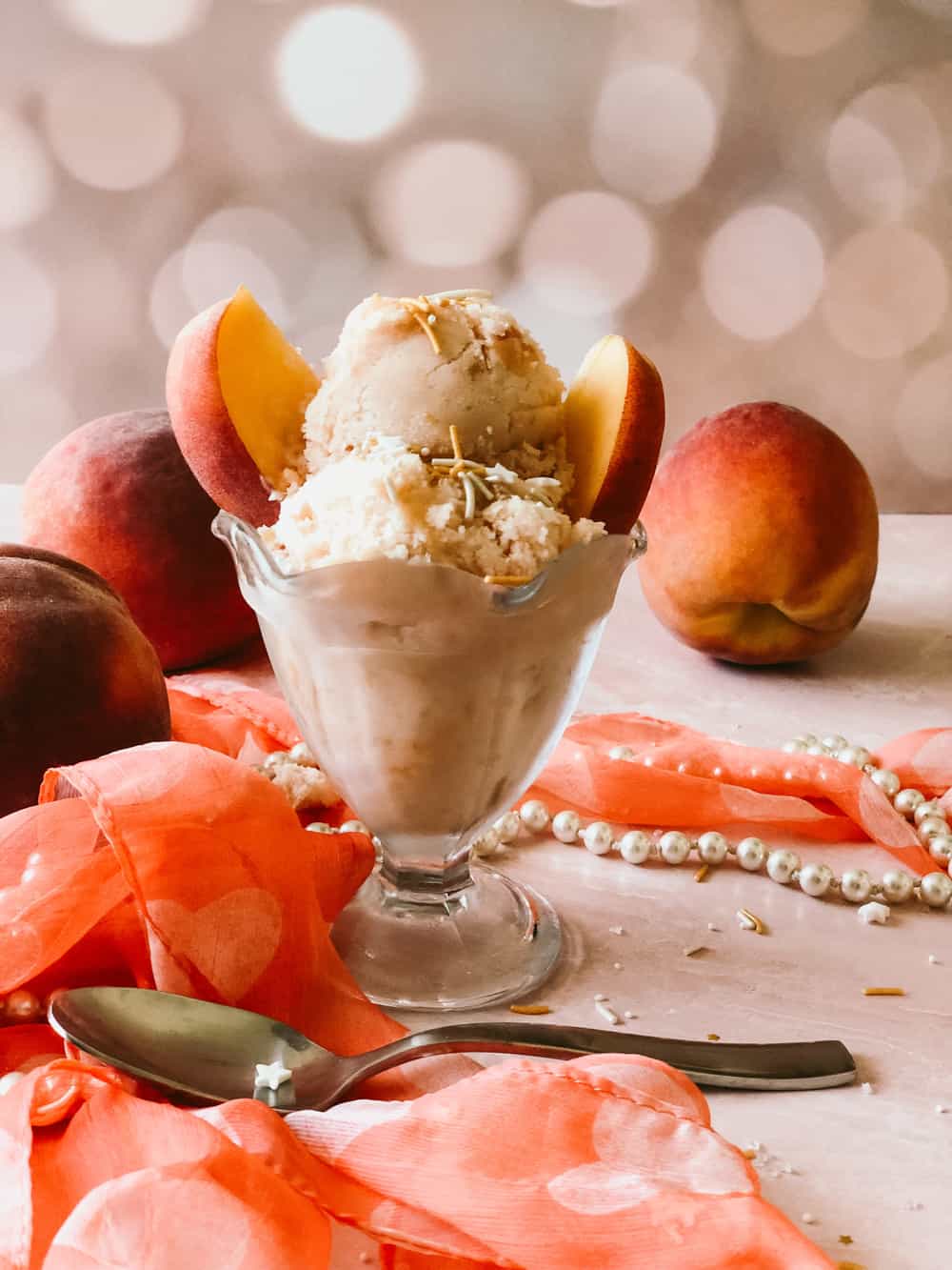 Peaches and cream ice cream in a glass dish with peaches sticking out of the top. a scarf, pearls, and spoon in the foreground. Peaches in the background.