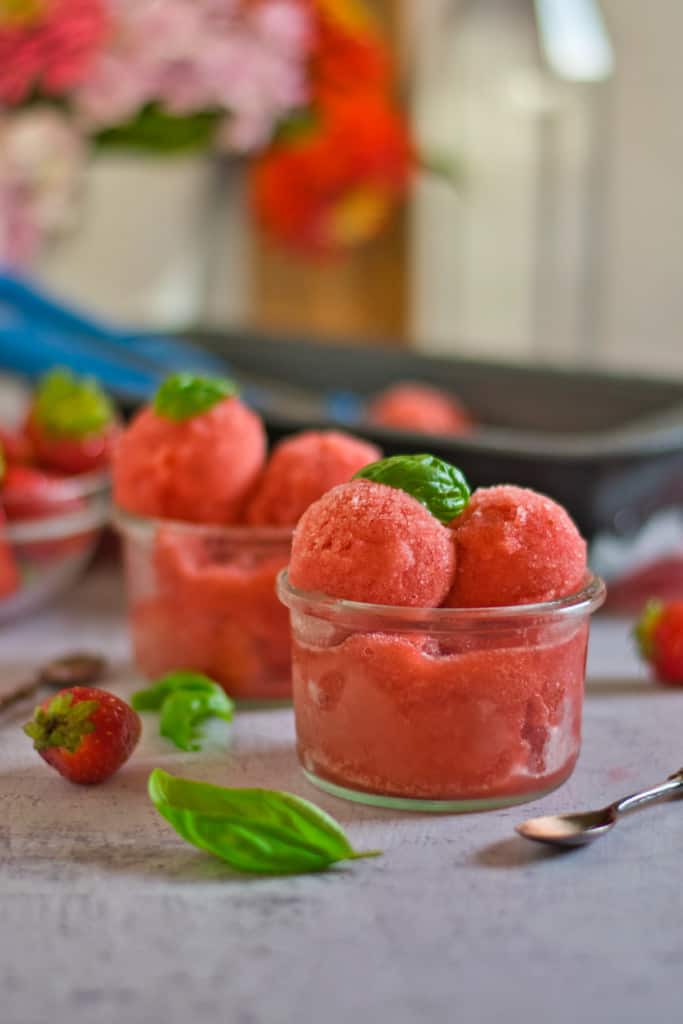 Scoops of strawberry basil sorbet in two glass ice cream dishes with the pan of sorbet in the background