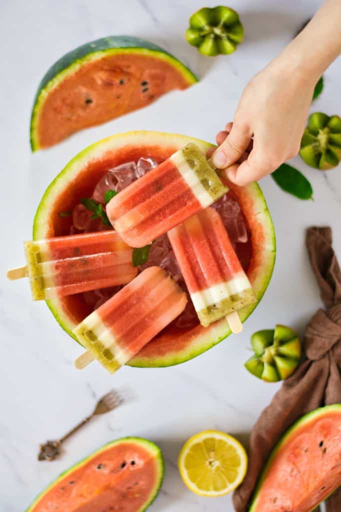 four watermelon kiwi popsicles in ice in a hollowed watermelon half with one held in hand
