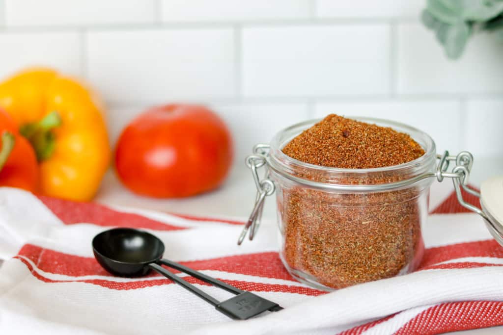 glass jar of homemade taco seasoning with peppers and tomato in background