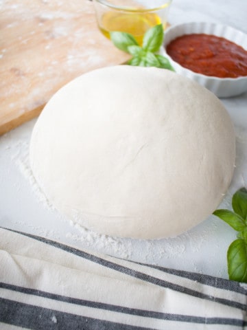 uncooked ball of pizza crust dough with basil leaves , pizza sauce, and olive oil in background
