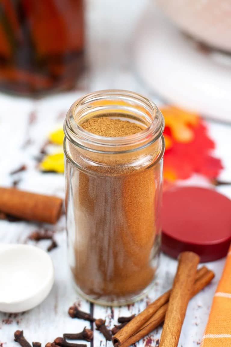 jar of pumpkin pie spice against seasonal background of cinnamon sticks, leaves, and whole cloves.