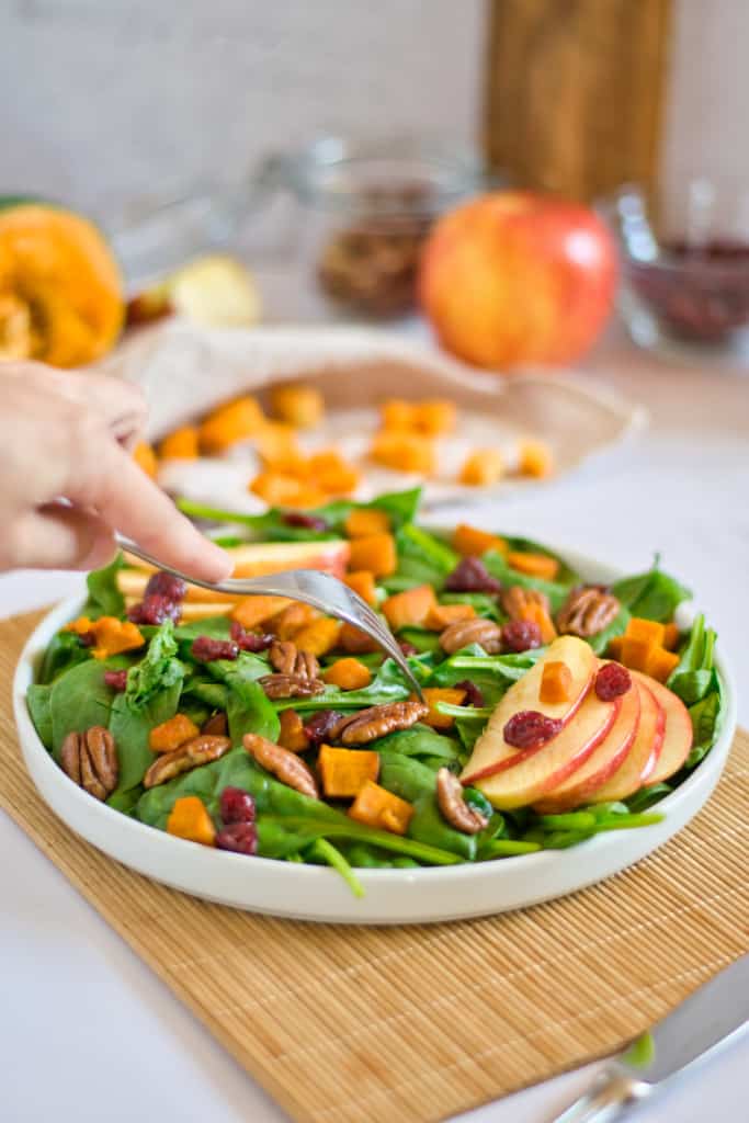 large serving of autumn salad with fork in hand, pecans, and pumpkin and background