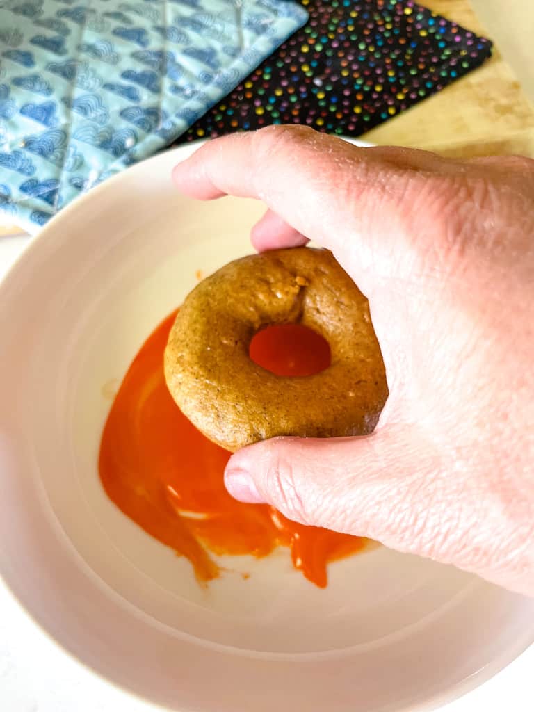 hand dipping baked donut into orange icing