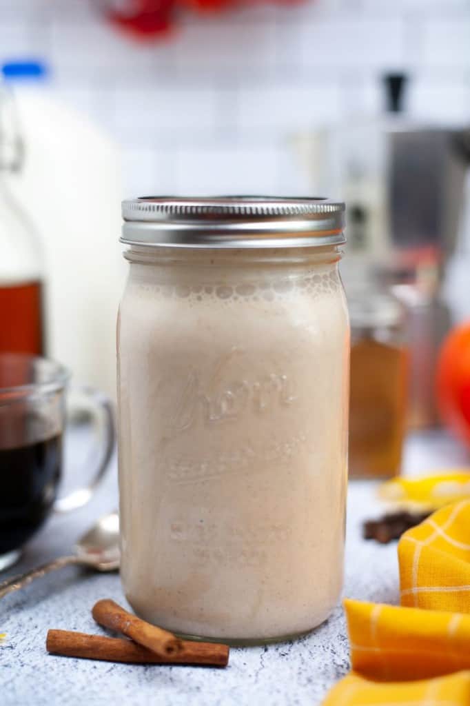 pumpkin spice creamer in mason jar with glass cup of coffee on the left and yellow napkin and orange pumpkin on the right