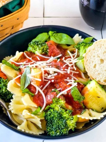 black bowl with pasta primavera topped with marinara sauce with red wine and bread basket in background