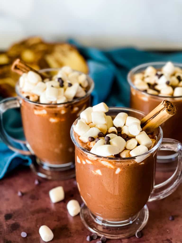 Three glass mugs of hot chocolate topped with marshmallows, mini chocolate chips, and cinnamon sticks. Slices of cake are in the background