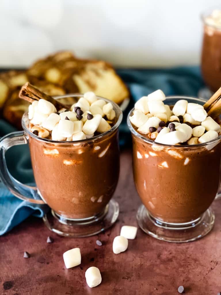 Three glass mugs of hot chocolate topped with marshmallows, mini chocolate chips, and cinnamon sticks. Slices of cake are in the background