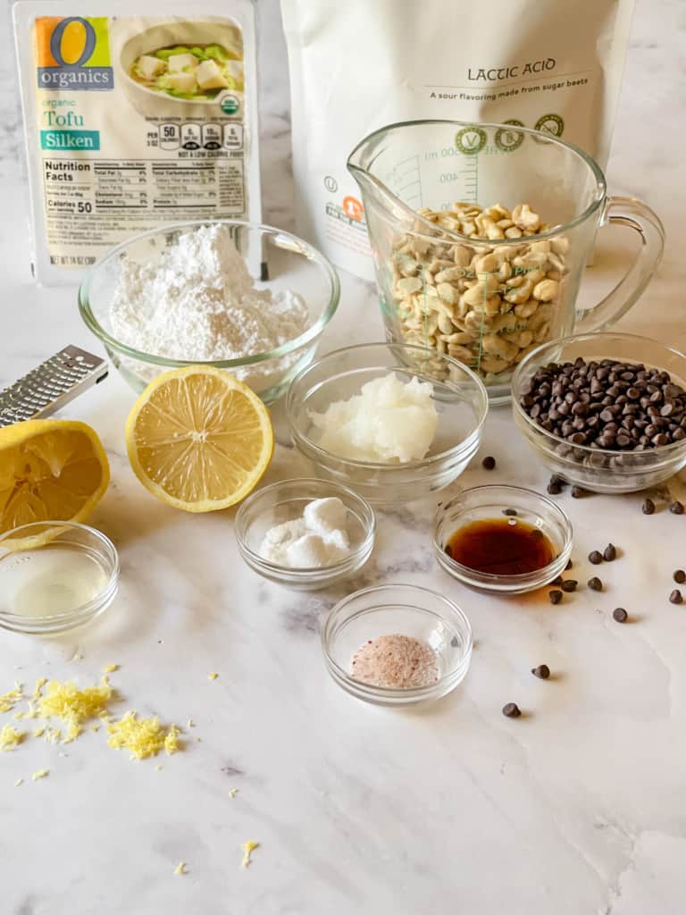 Ingredients needed for cheesecake cannoli dip including silken tofu, lactic acid, cashews, powdered sugar, lemon and zest, coconut oil, vanilla, salt, and mini chocolate chips