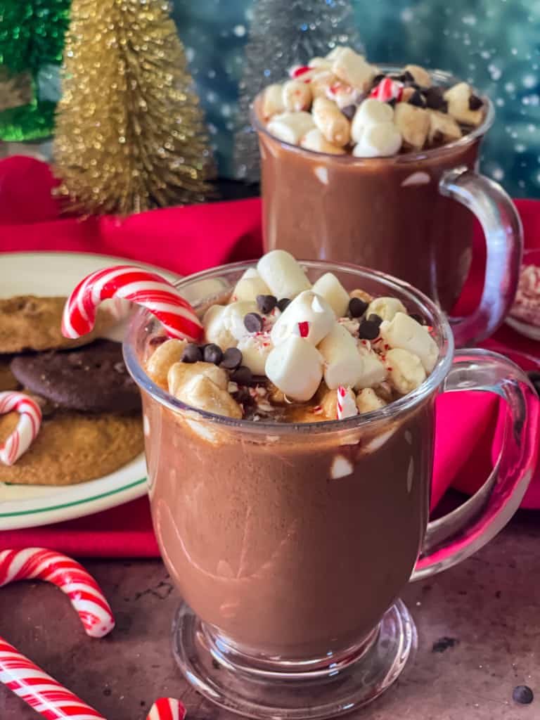 Two glass mugs of peppermint hot chocolate topped with marshmallows, mini chocolate chips, and candy canes. A plate of cookies are in the background along with festive sparkly trees