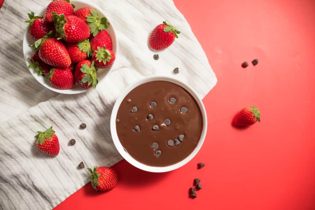 chocolate dip in white bowl with chocolate chips sprinkled on the top and a bowl of strawberries with some chocolate chips and strawberries surrounds the bowls