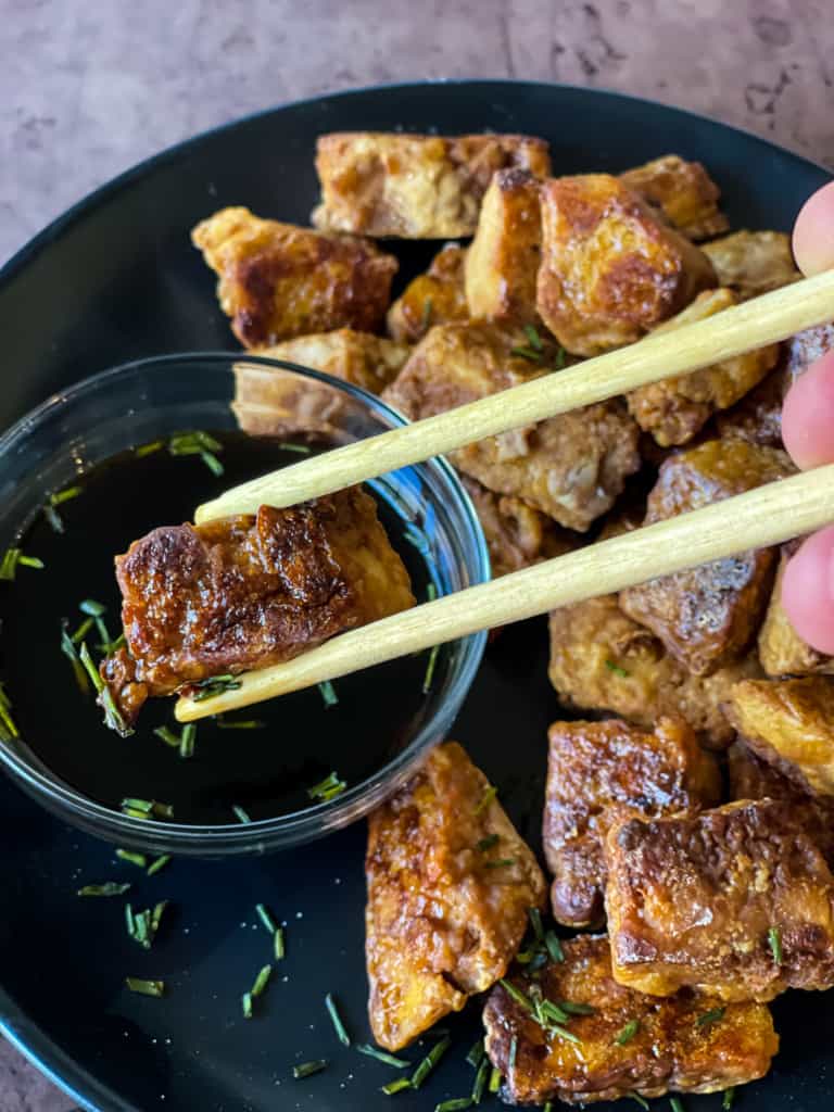 Wooden tongs dipping a piece of baked tofu into a soy sauce dipping sauce