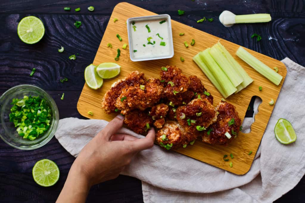 tray of cooked buffalo cauliflower wings with a hand in photo placing a wing