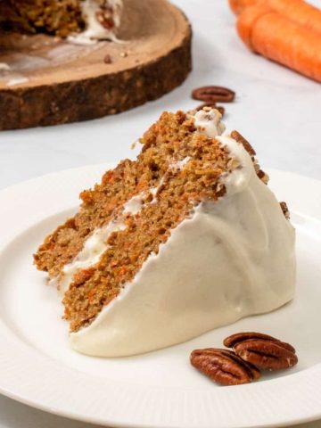 Slice of vegan carrot cake with cake and carrots in background