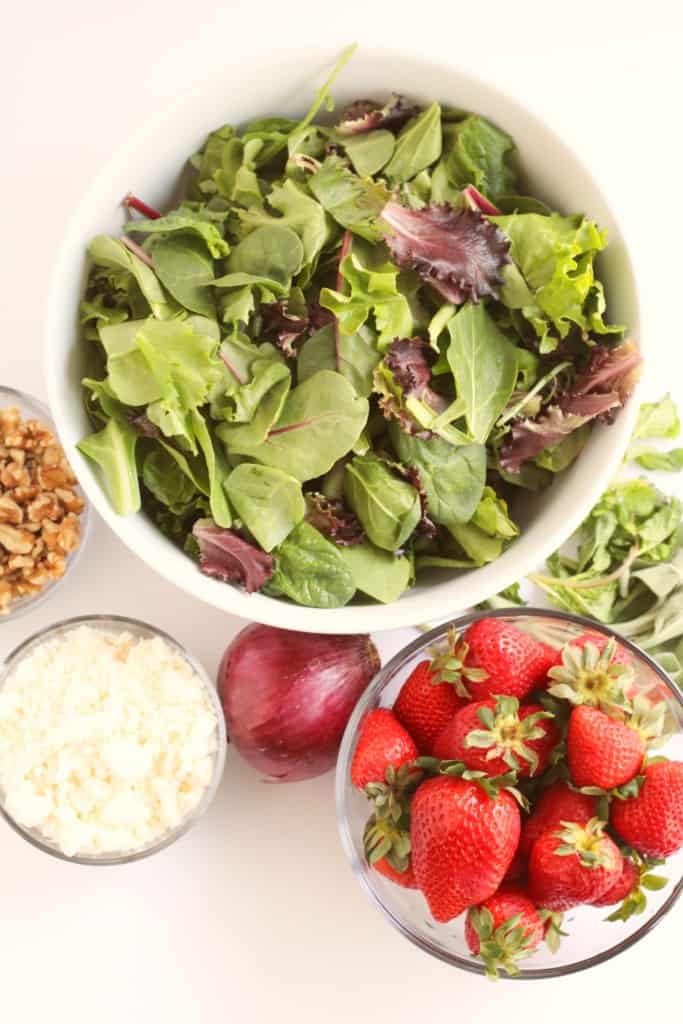 getting ready to assemble the strawberry walnut salad with a big bowl of spring mix lettuce, walnuts, vegan feta, shallot, and strawberries next to some sage and mint leaves