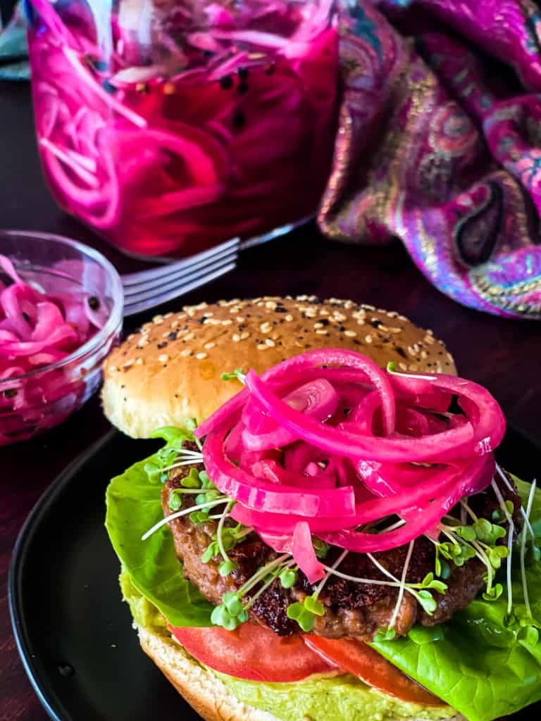 burger with pink onions with big jar and two small bowls with pink onions with fork in background