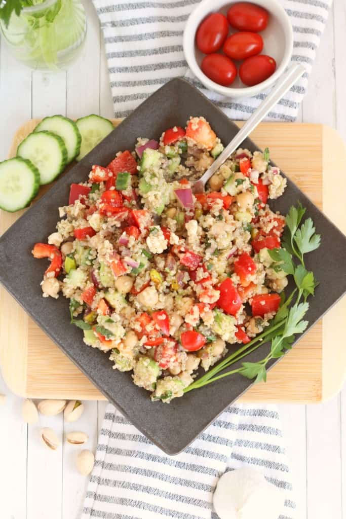 prepared quinoa salad with chickpeas on a dark slate plate with sliced cucumber, and cherry tomatoes in background with fork in salad