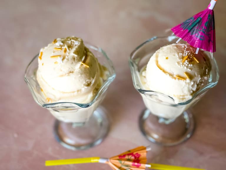 two glass ice cream dishes with pina colada ice cream in front of a window with two umbrella straws in foreground