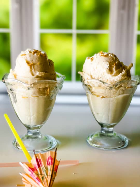 two glass ice cream dishes with pina colada ice cream in front of a window with two umbrella straws in foreground