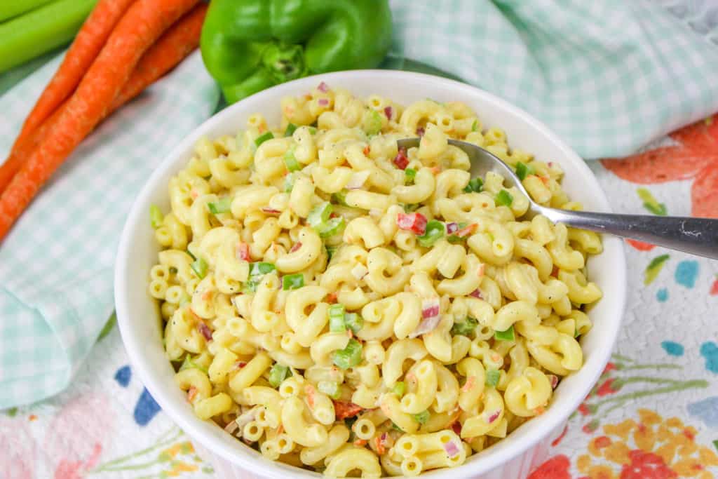 bowl of vegan macaroni salad with carrots, green pepper, and celery in the background with a spoon in the dish