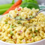 bowl of vegan macaroni salad with carrots, green pepper, and celery in the background with a spoon taking a portion