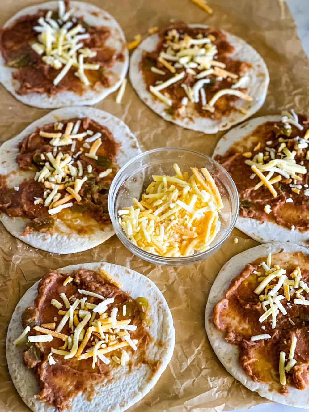 six small tortillas with refried beans a cheese in the center of each with a bowl of shredded cheese in the center of the tacos