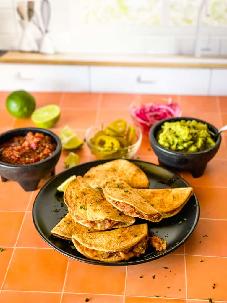 plate of tiny tacos on the kitchen counter with salsa, guacamole, lime, jalapeno, and pickled onions in the background