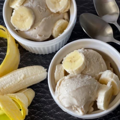 two white bowls of frozen yogurt with banana sliced on top and on the side with two spoons